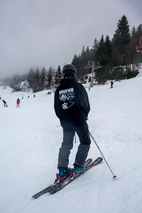 A skier standing with poles on a piste, seen from behind.