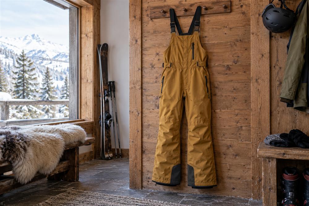 Snow bib trousers with a chest panel and adjustable shoulder straps hanging on a wooden wall