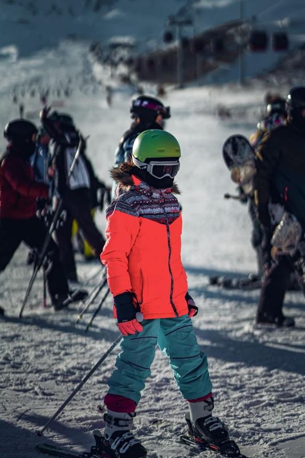 A child stands on the piste wearing ski clothing with skis and poles.