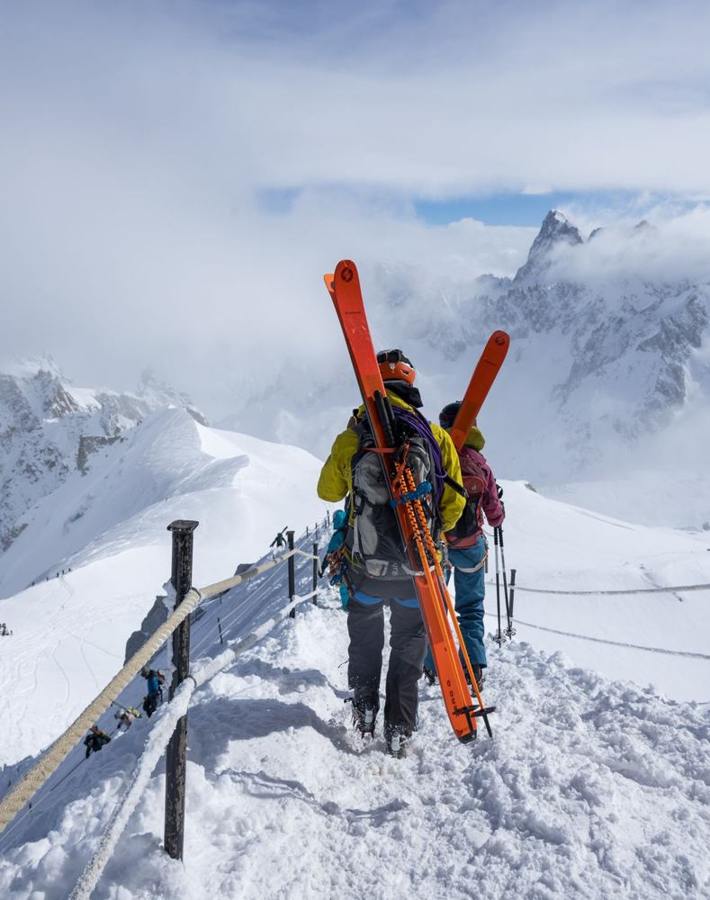 Two people carry winter sports equipment along a snowy mountain path.