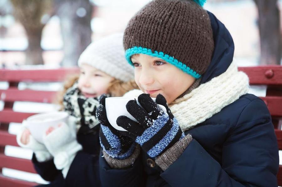 Two children in winter clothing drink from cups on a bench.