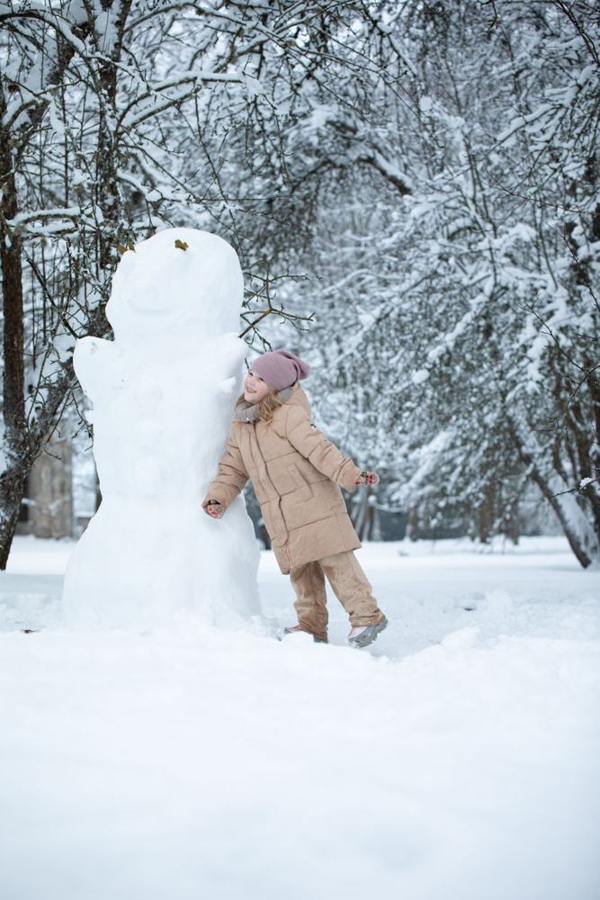 A child stands beside a large snowman in a snowy park.