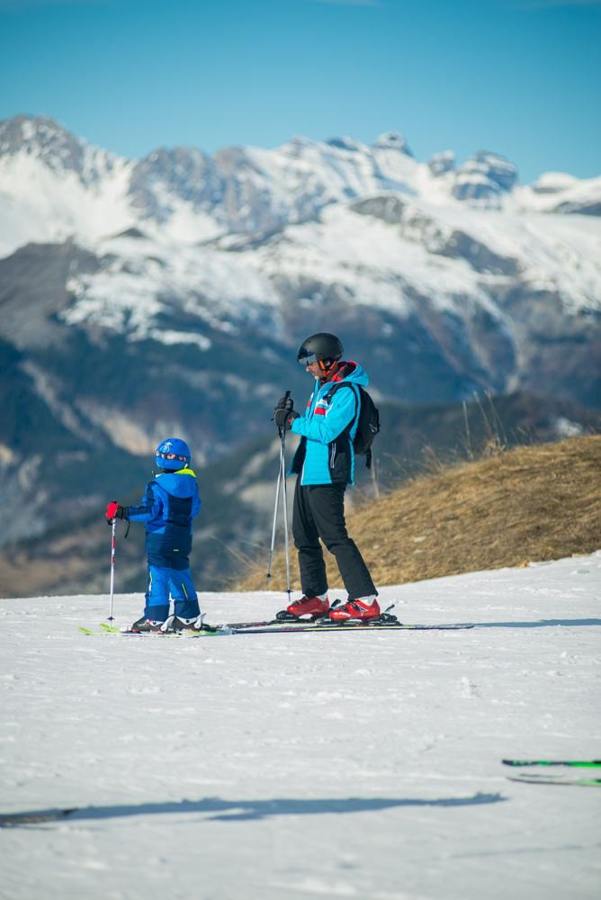 An adult and a child stand on skis on a piste below mountains.
