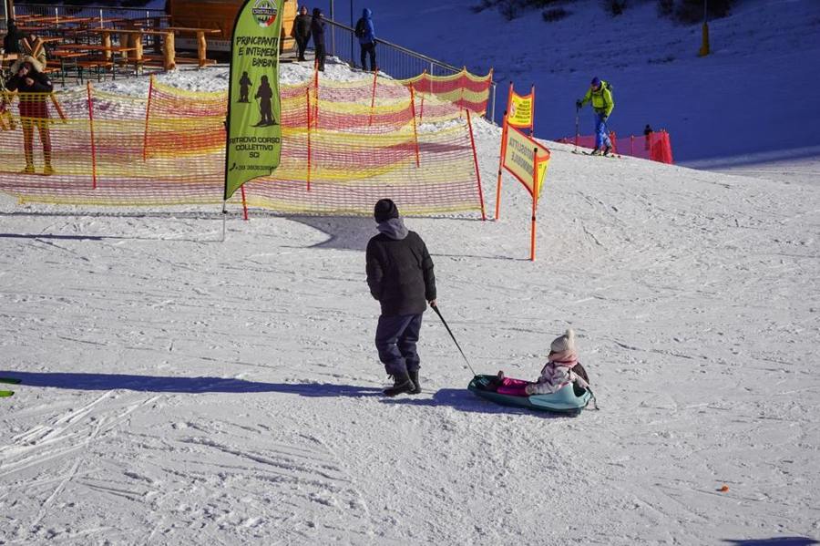 An adult pulls a child on a sledge beside a ski piste.