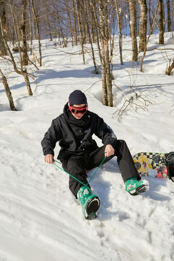 A snowboarder sits on snow and adjusts snowboard boots.
