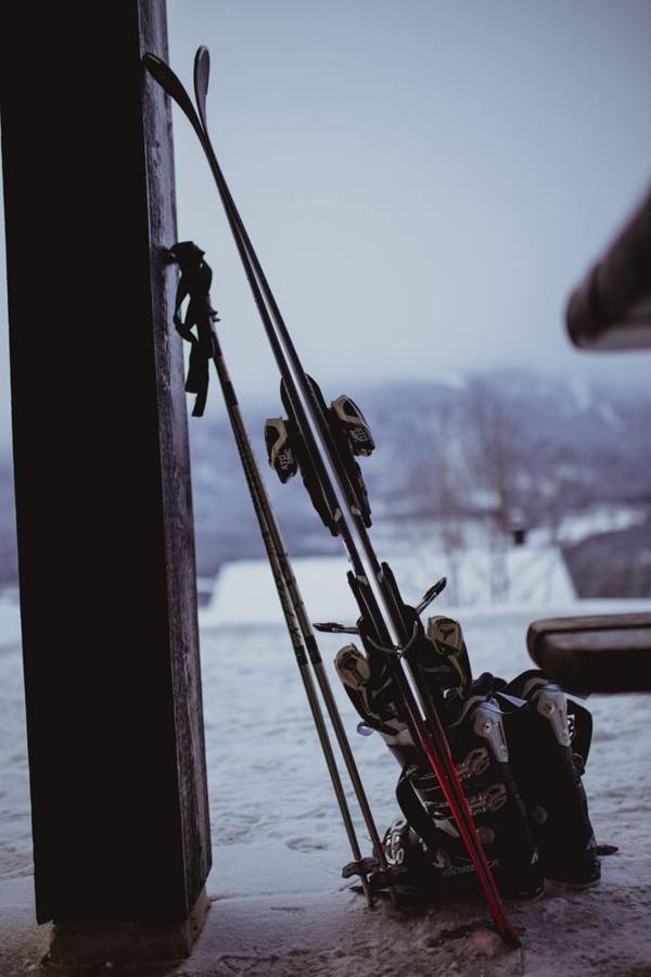 Skis, poles and ski boots stand in a sheltered area beside a wooden wall.